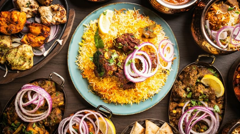 A vibrant spread of Indian dishes, including biryani, curries, and grilled meats, garnished with onions and lemon.