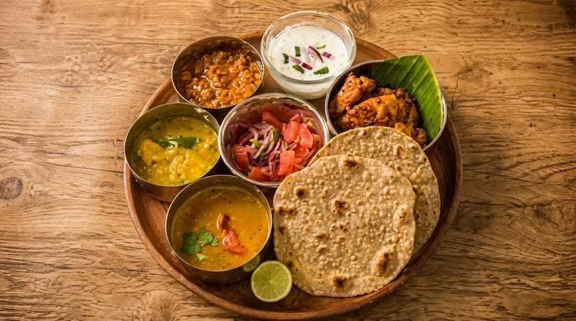 A wooden platter with various Indian dishes, including curries, flatbread, raita, and salad.