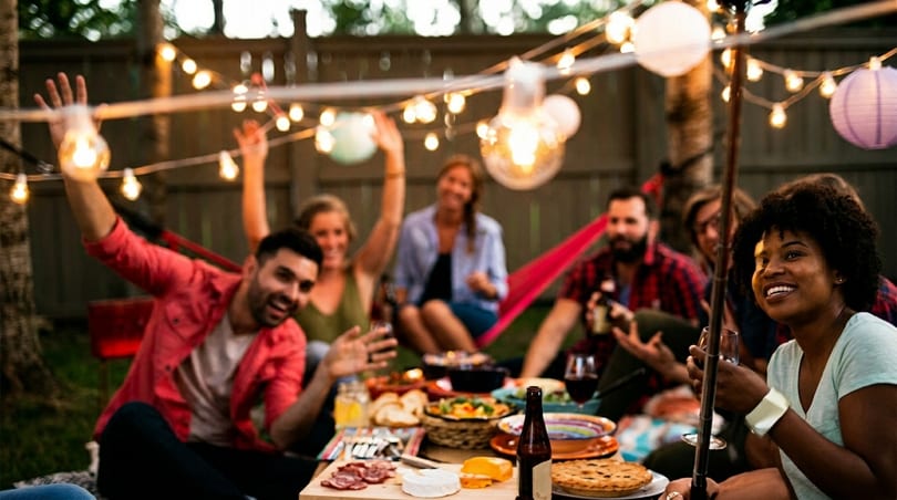 Friends enjoying an outdoor evening party with string lights, food, and drinks.