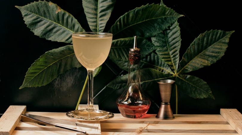 A cocktail glass with a light-colored drink, a bottle of red liquid, and a jigger on a wooden surface.