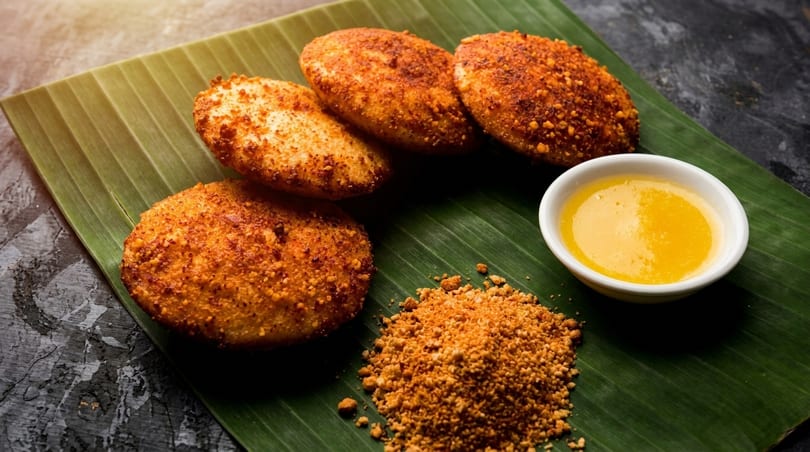 Four golden-brown fried idlis, a bowl of ghee, and chili powder on a banana leaf.