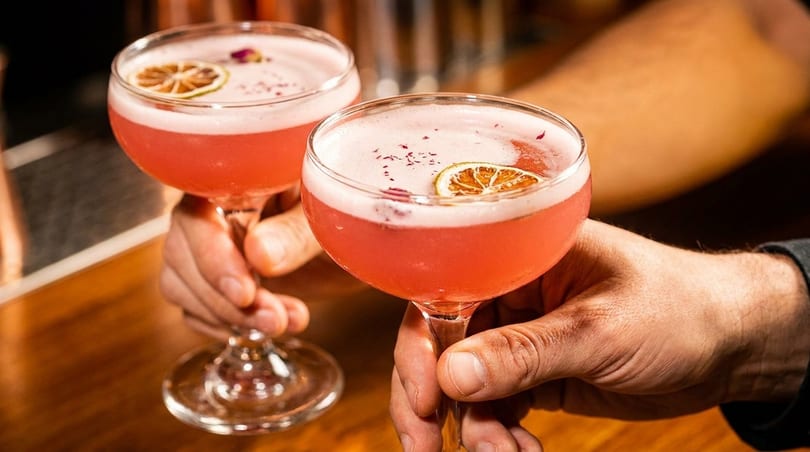 Two hands holding pink cocktails with lime slices and rose petals on a wooden bar.